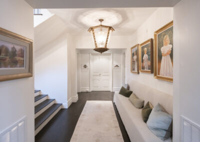 A large chandelier lights a white hallway with portraits on the wall within a home